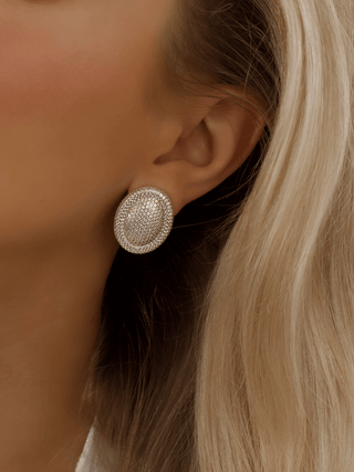 Gold plated Gigi pavé oval stud earrings worn on model. Covered in shimmering clear CZ stones with a sculptural oval shape for high-impact glamour.