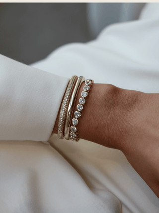 Gold heart tennis bracelet worn with polished and pavé bangles, styled on wrist in natural light for an elegant, high-impact stack