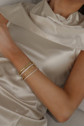 Gold plated pavé slim Christy bangle stacked on model’s wrist with gold cuffs and CZ bracelets. Elegant jewellery styling in satin outfit.