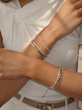 Plain gold plated slim Christy bangle styled with pavé Christy bangle and Micro Hydrangea Bracelet on model in white outfit. Stacked gold bracelets for summer styling and special occasions.