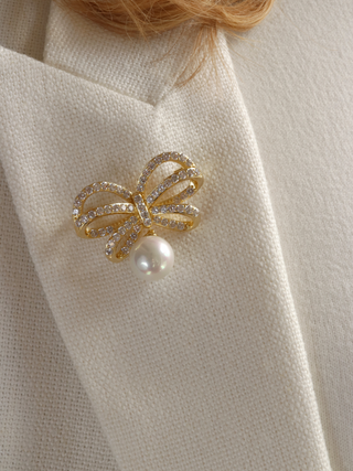 Meredith bow-shaped brooch adorned with sparkling crystals and a shell pearl, styled on the lapel of a model’s white coat, perfect for formal events and elegant summer outfits.