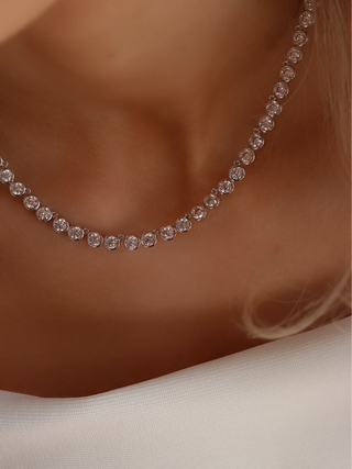 Serena tennis necklace featuring a continuous line of round CZ stones bezel-set in white gold plating, shown in a dynamic close-up as the model leans forward, highlighting the necklace's sophisticated sparkle and fluid elegance.