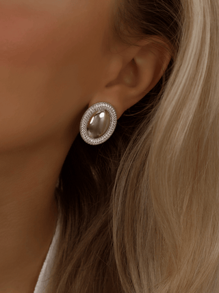 White gold plated Gigi semi pavé oval stud earrings with clear CZ stones, worn on model. Bold sculptural design with pavé crystal border for modern elegance.