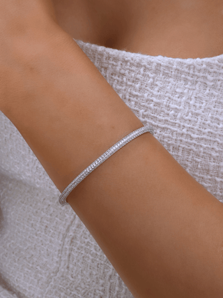White gold plated pavé slim Christy bangle worn on wrist against white textured fabric. Dainty half-set CZ bracelet with elegant sparkle for everyday styling.