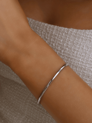 White gold plated slim Christy bangle worn on wrist against white textured fabric. Minimalist polished bangle with smooth finish, ideal for everyday stacking.
