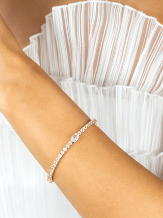 Gold plated tennis bracelet featuring central oval clear CZ stone, worn on wrist with white pleated dress