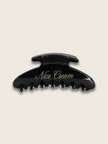 Flatlay of a shiny black claw hair clip, with the brand name written on the side, displayed on a plain white background