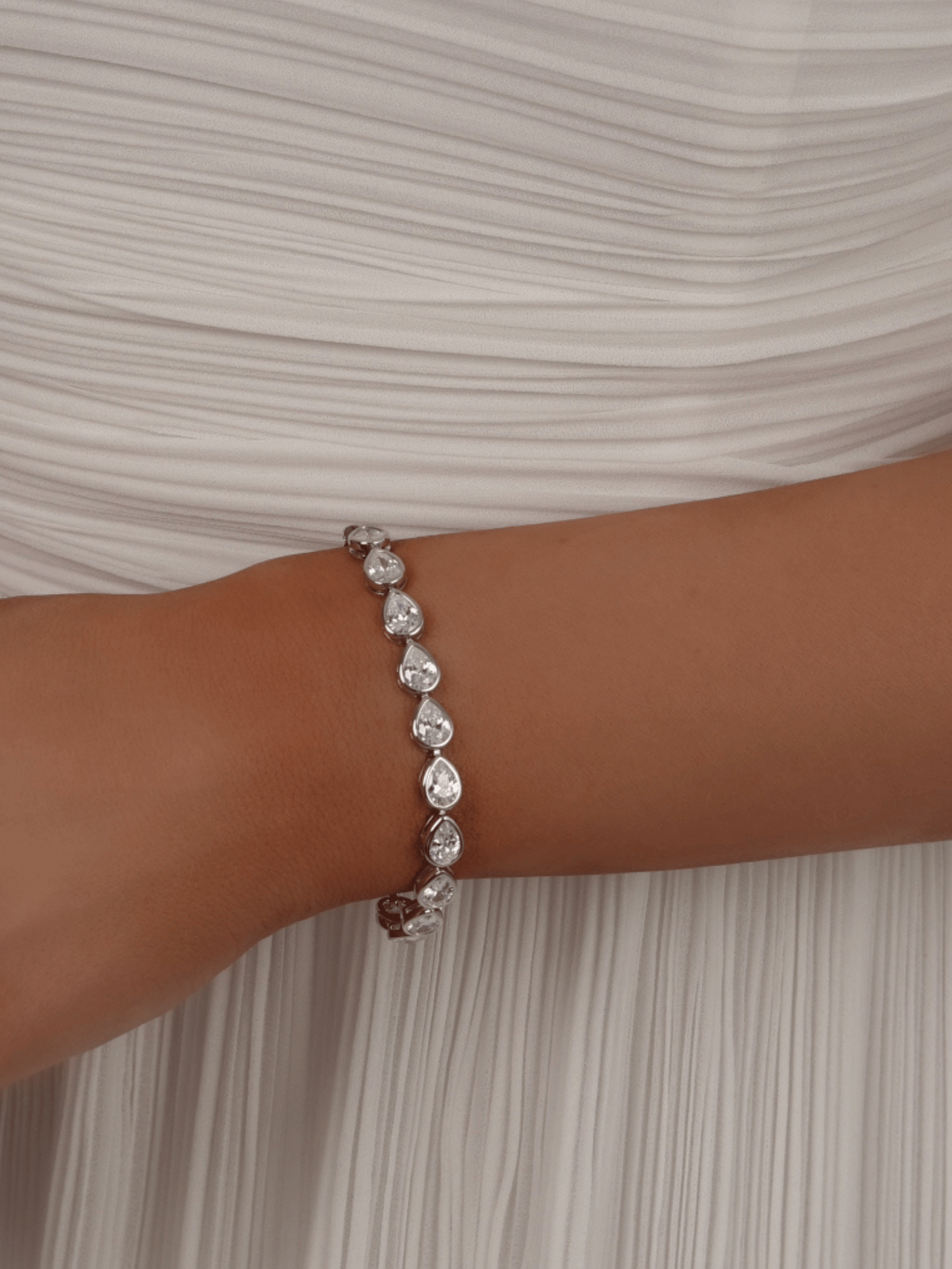 Constance pear-shaped CZ tennis bracelet in white gold plating, worn on a model's wrist with a white dress in the background, showcasing elegant bridal or formal jewellery styling.