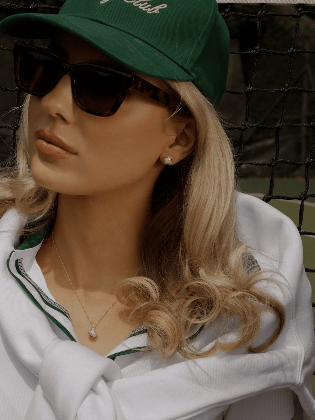 Model wearing gold pavé tennis ball stud earrings with matching necklace, styled in tennis whites and green cap