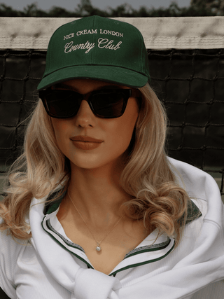 Model wearing gold plated tennis ball necklace with pavé CZ pendant, styled with Nice Cream London cap and tennis whites