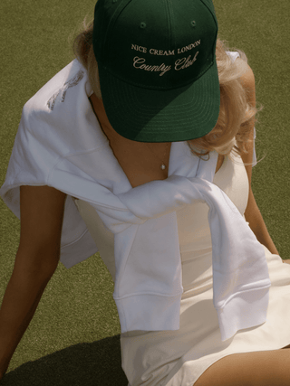 Overhead shot of model on tennis court wearing gold pavé tennis ball necklace and Nice Cream London Country Club cap