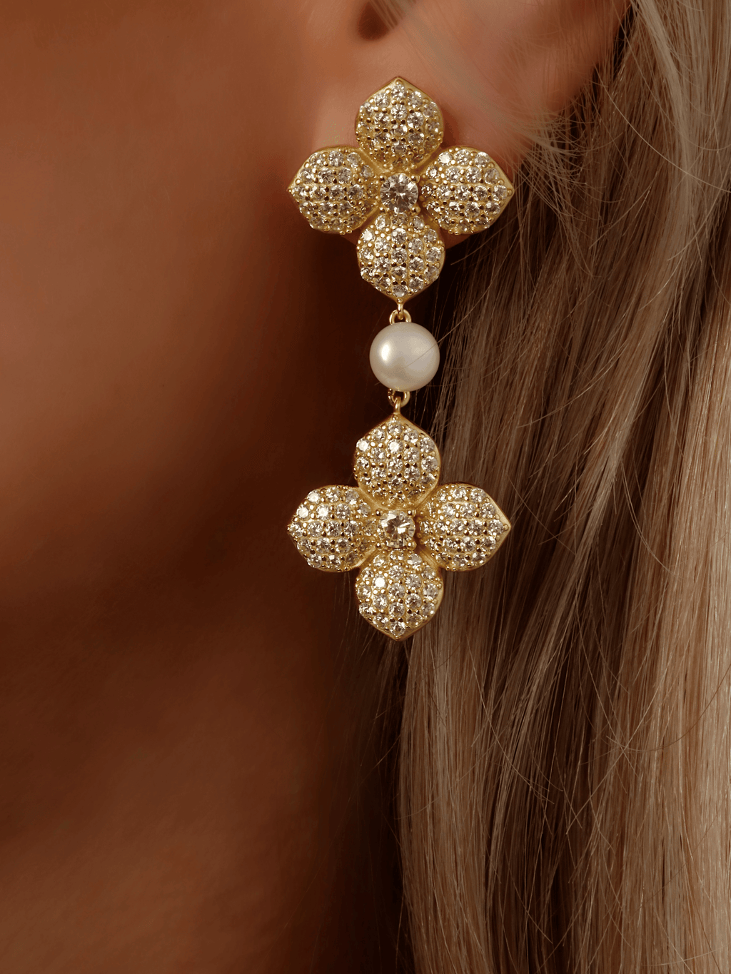 Close-up of model wearing gold plated dangle earring with two hydrangea flowers, yellow cubic zirconia stones and freshwater pearl. Sparkly floral statement earring for weddings and special occasions.