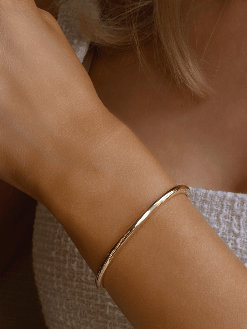 Gold plated slim Christy bangle worn on wrist against white textured fabric. Minimalist polished gold bracelet with sleek finish, perfect for everyday stacking.