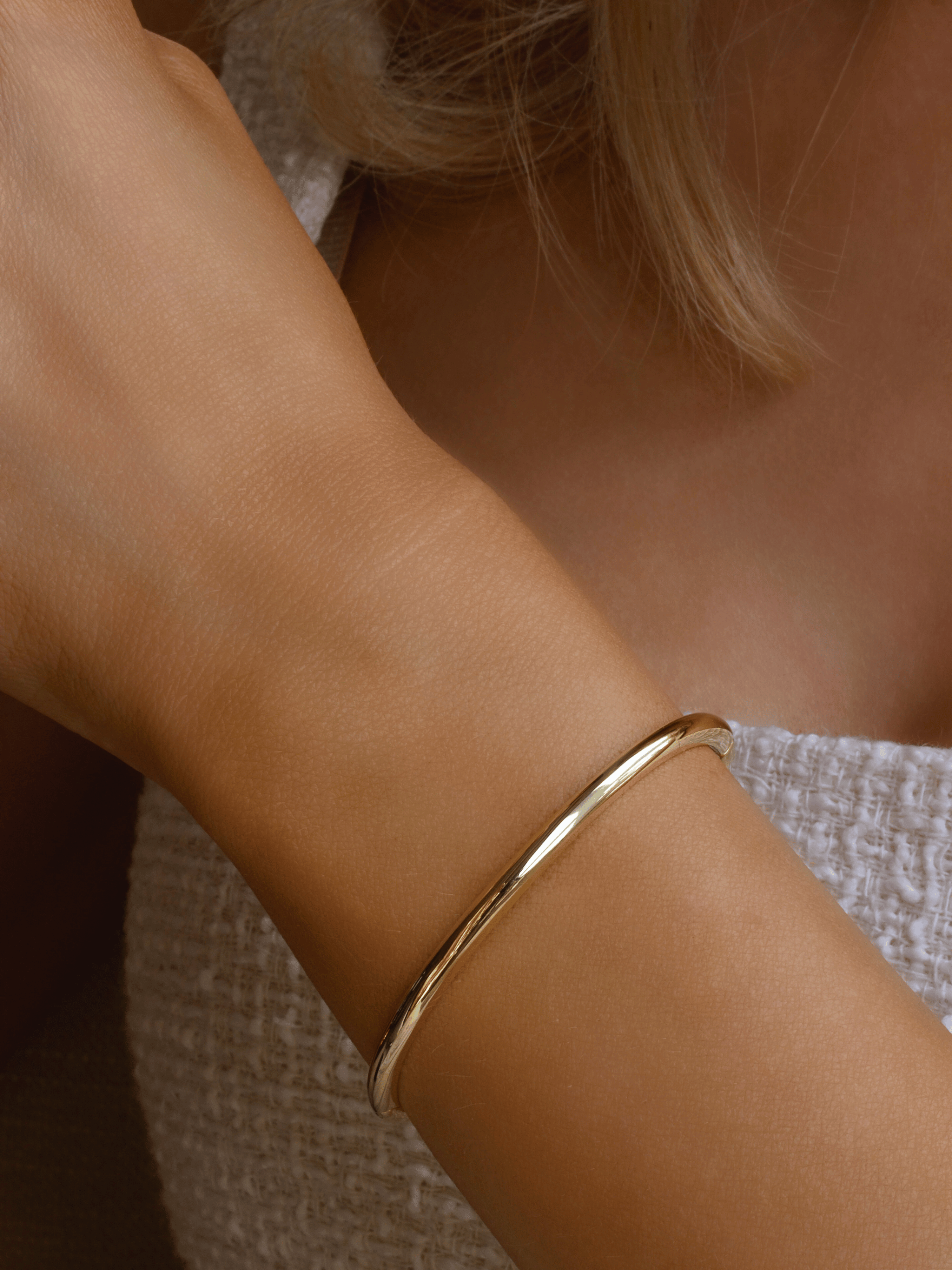Gold plated slim Christy bangle worn on wrist against white textured fabric. Minimalist polished gold bracelet with sleek finish, perfect for everyday stacking.