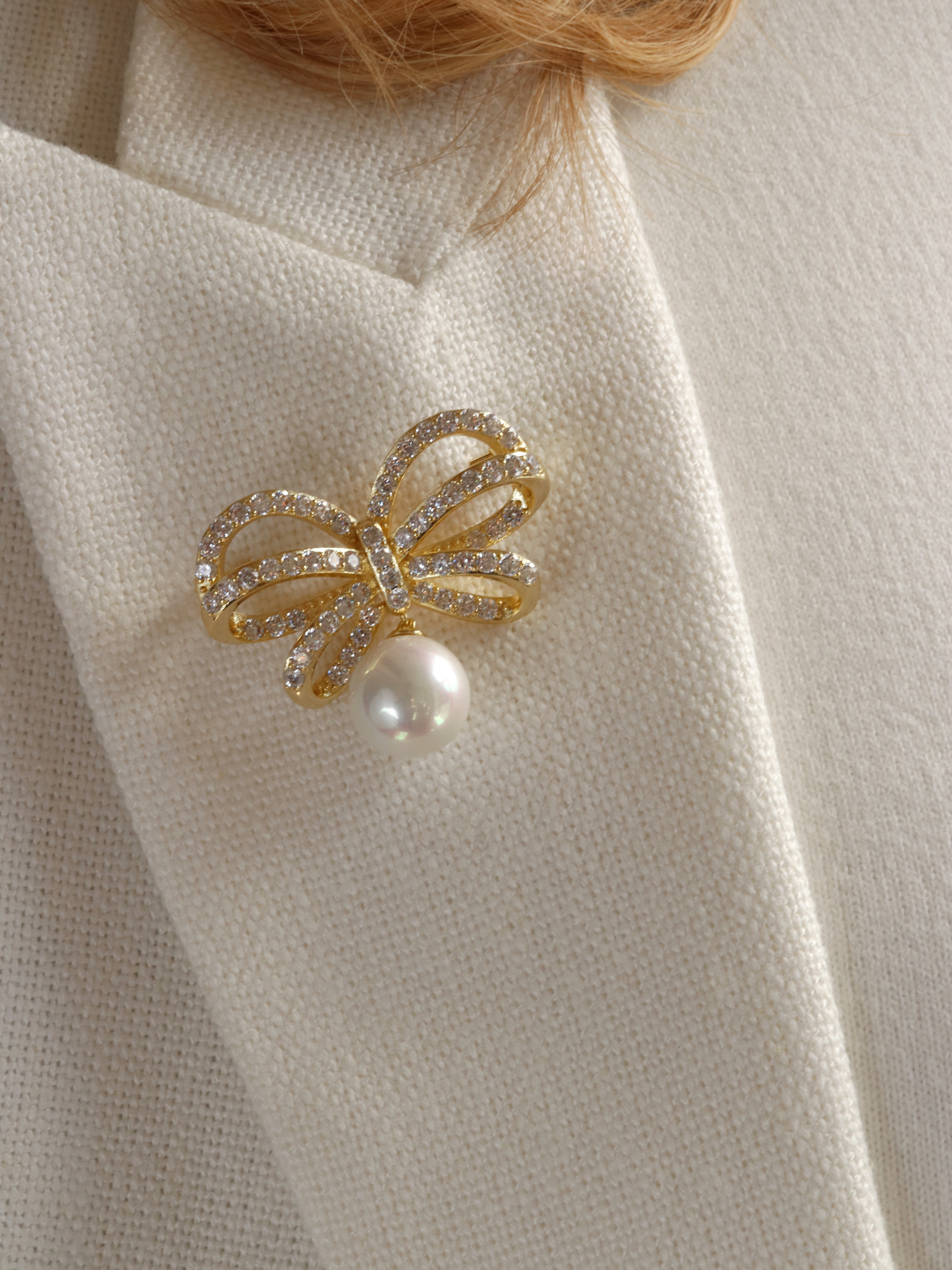 Meredith bow-shaped brooch adorned with sparkling crystals and a shell pearl, styled on the lapel of a model’s white coat, perfect for formal events and elegant summer outfits.