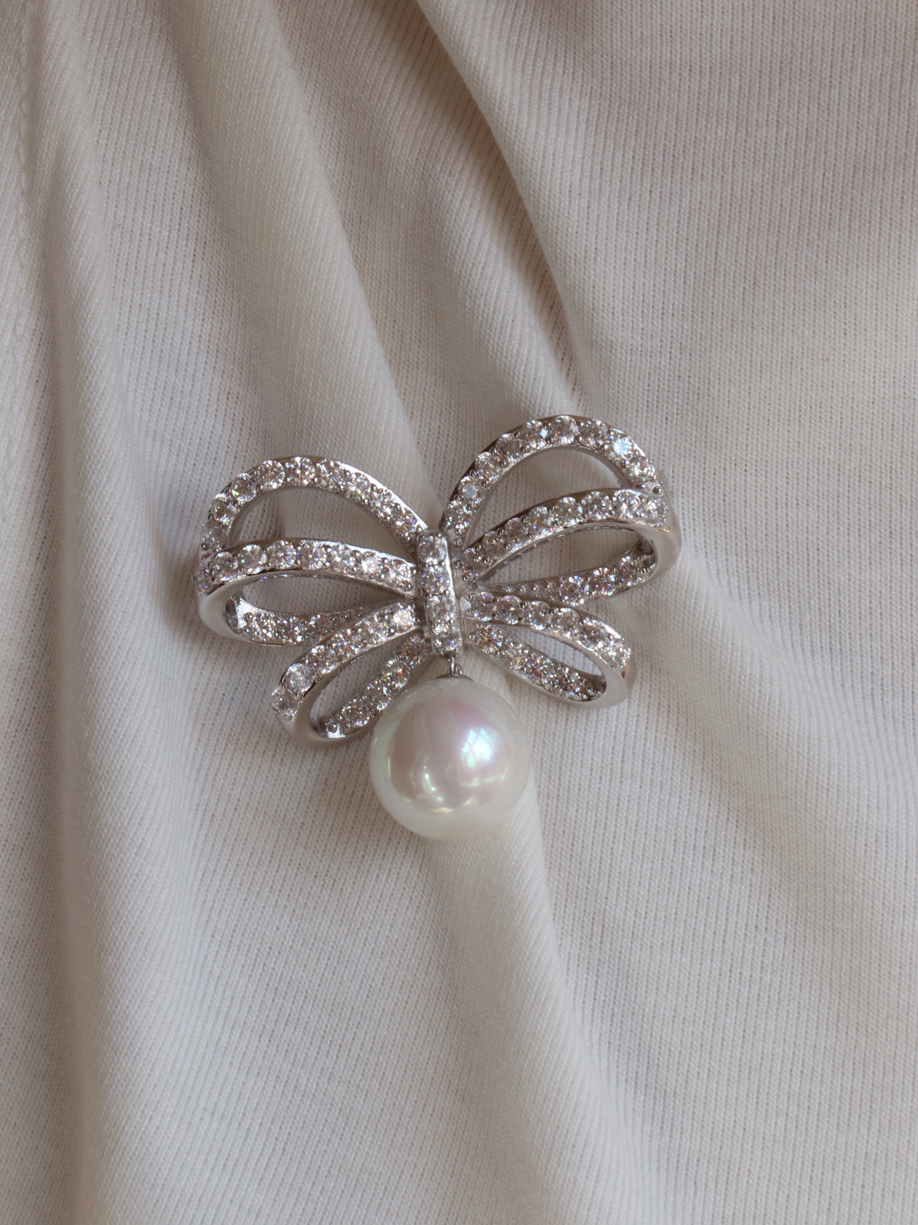 Close-up of the Meredith bow brooch in white gold plating, featuring sparkling crystals and a shell pearl, styled on a white cotton T-shirt for an elegant and versatile summer look.