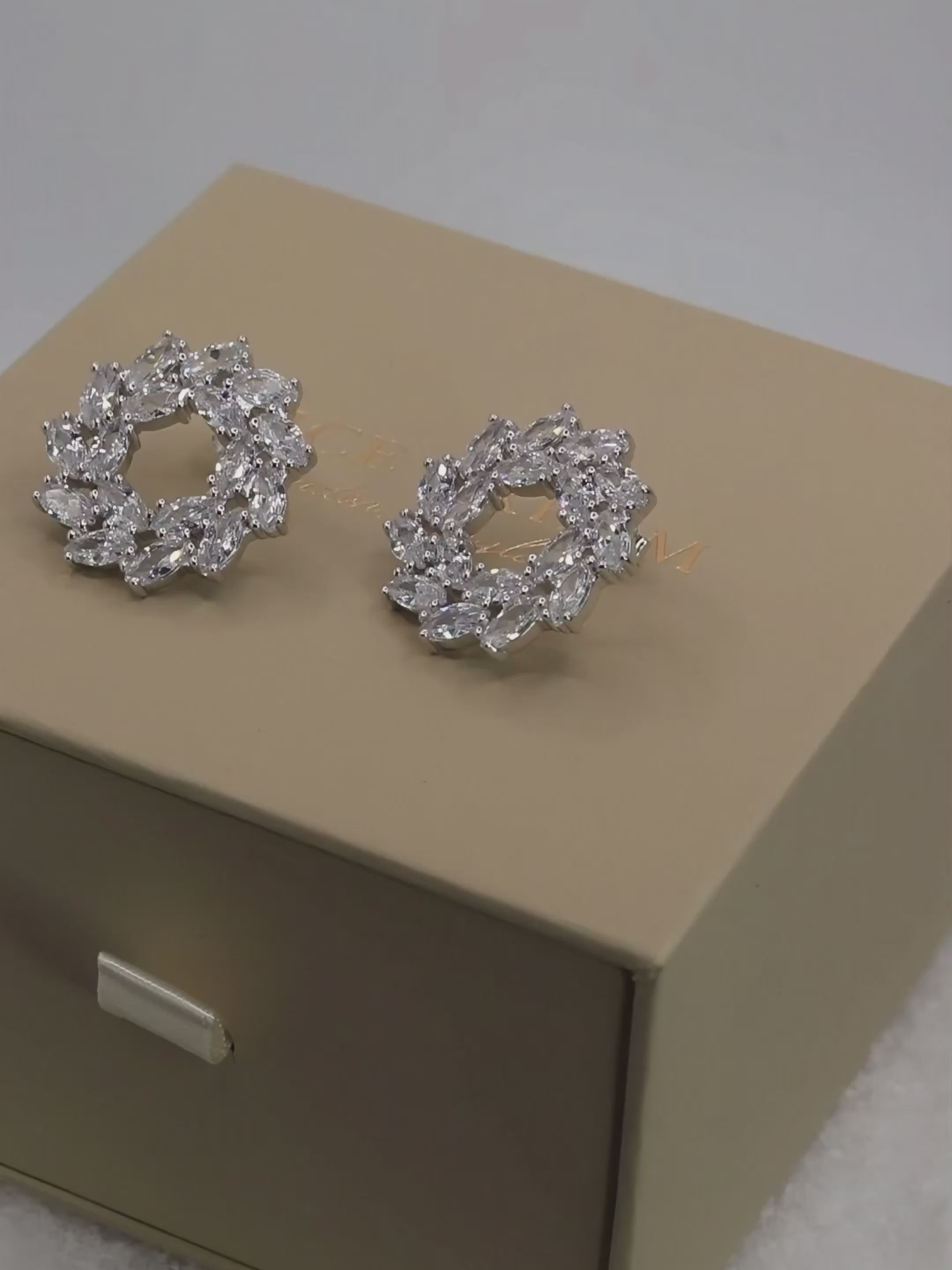 White gold Amanda stud earrings displayed on branded packaging, rotating on a neutral background to highlight the sparkle and intricate stone arrangement