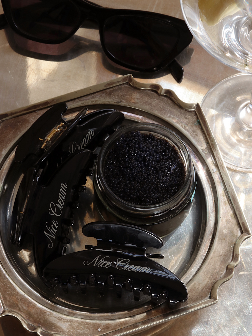 Overhead view of three black claw hair clips in the colour Caviar arranged on a silver tray with a glass pot of caviar and a pair of black sunglasses