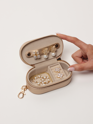 Small champagne jewellery case open with earrings, ring slots and pearl pieces arranged inside, shown with a hand.