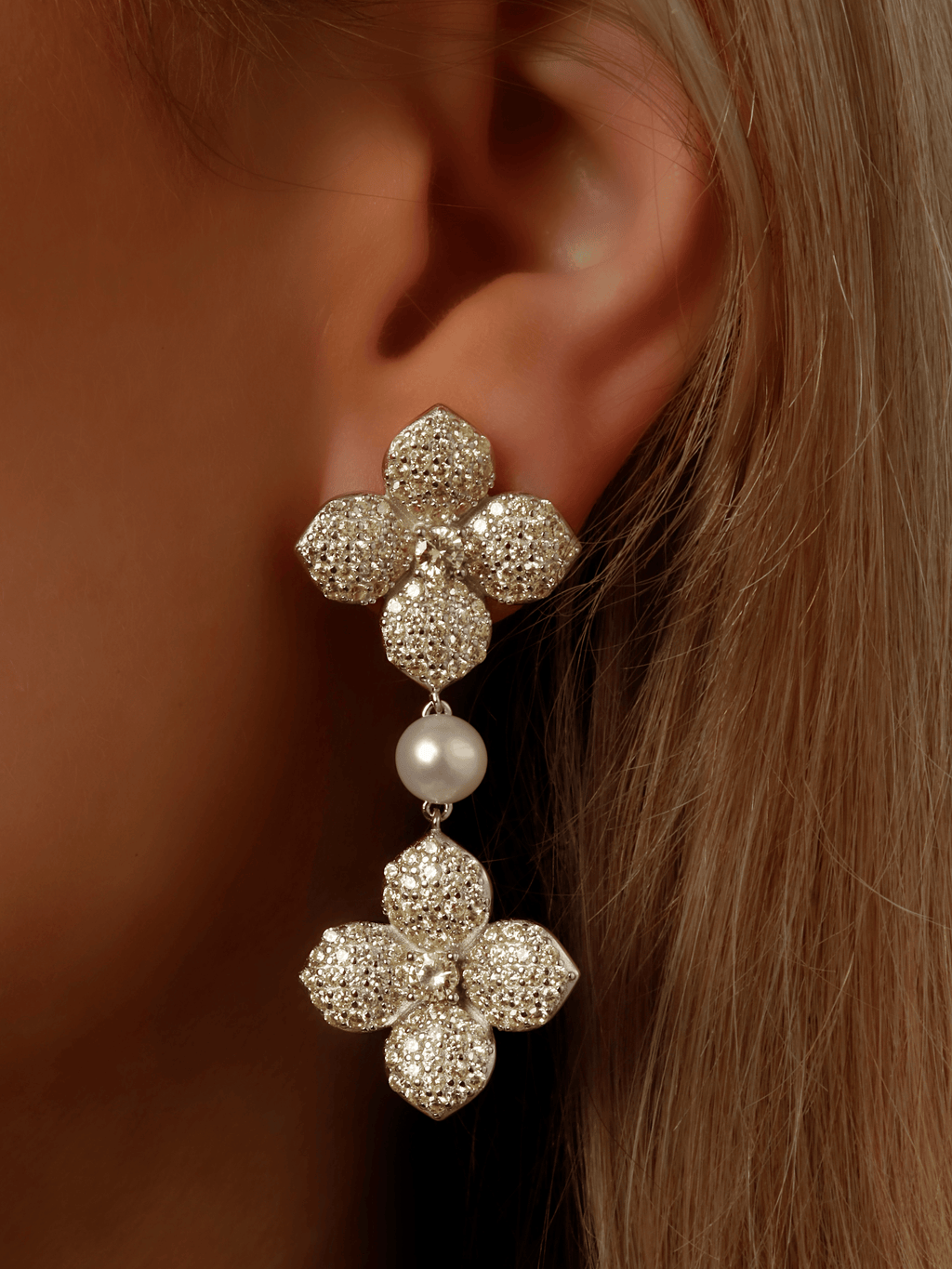 Close-up of model wearing white gold plated dangle earring with two hydrangea flowers, yellow cubic zirconia stones and freshwater pearl. Elegant sparkly statement earring ideal for bridal looks and evening wear.