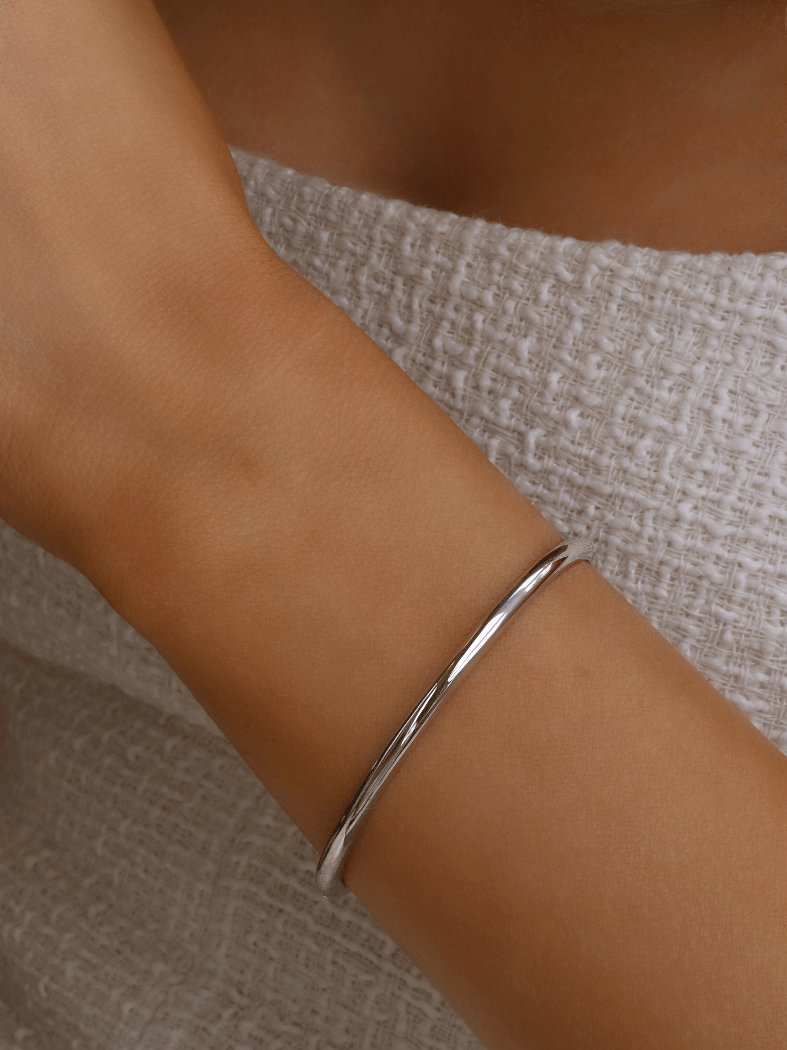 White gold plated slim Christy bangle worn on wrist against white textured fabric. Minimalist polished bangle with smooth finish, ideal for everyday stacking.